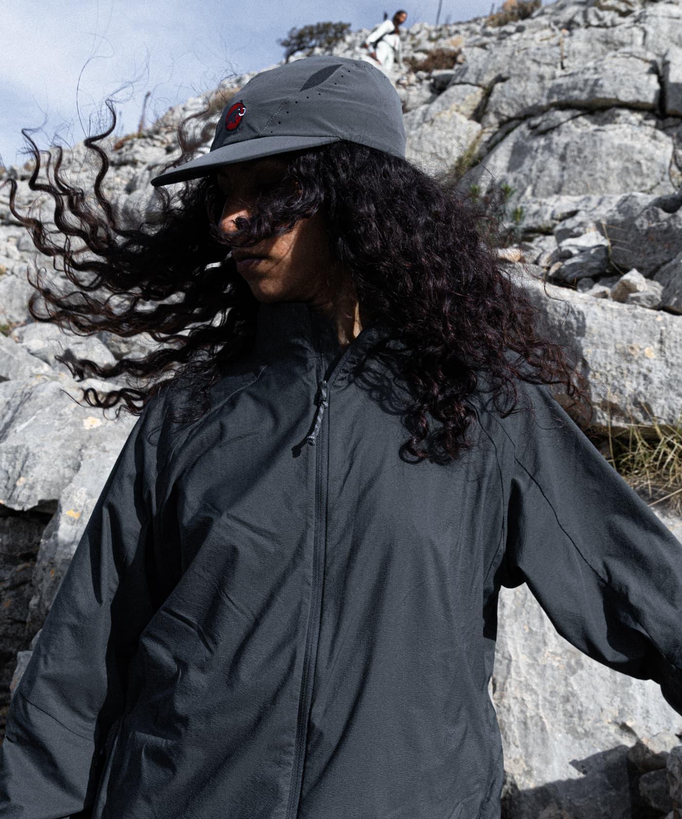 Person in a Mammut black jacket and cap with long curly hair standing by rocky mountain terrain on a windy day.