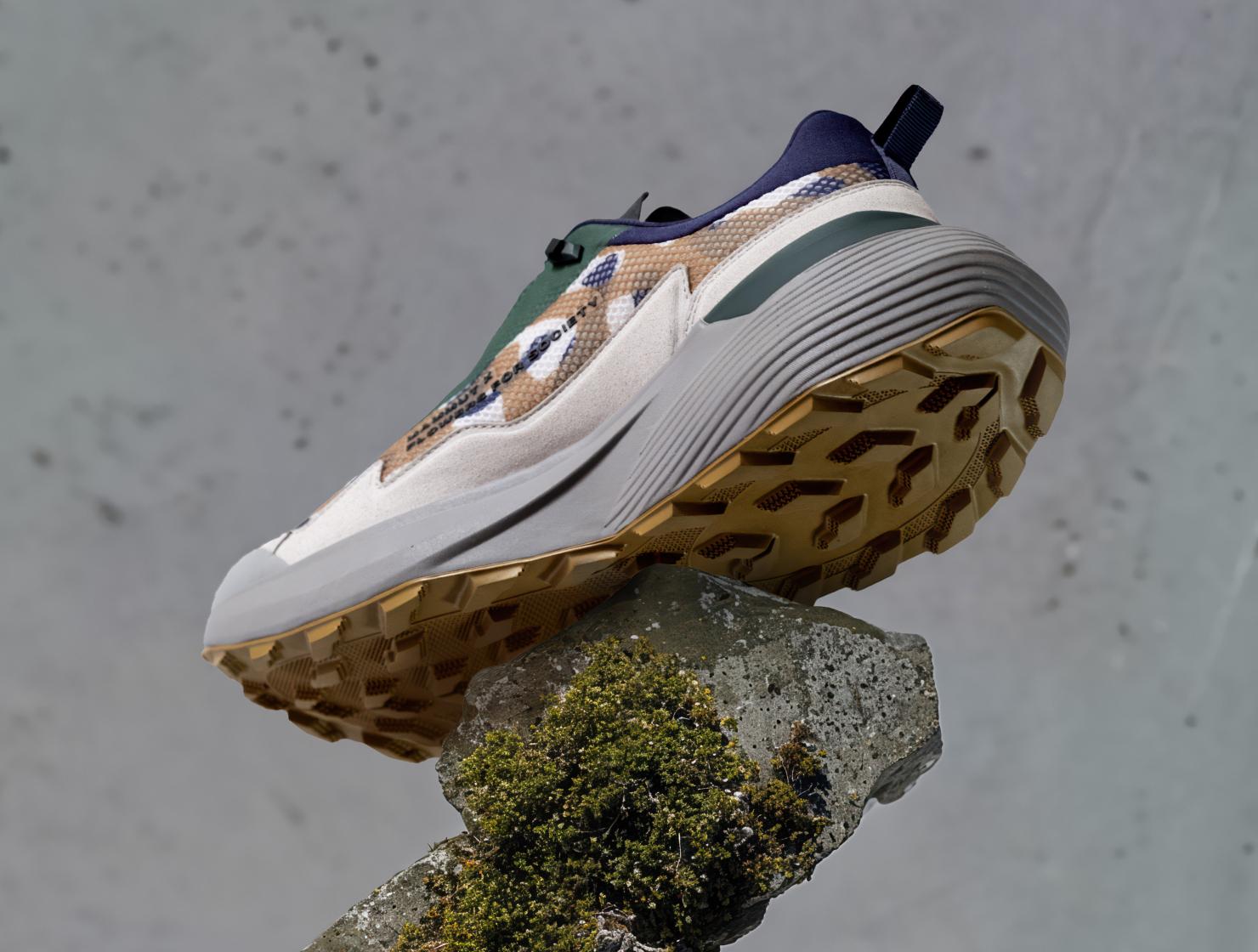 Beige and green Mammut trail sneaker with a chunky sole, balanced on a mossy rock against a gray background.
