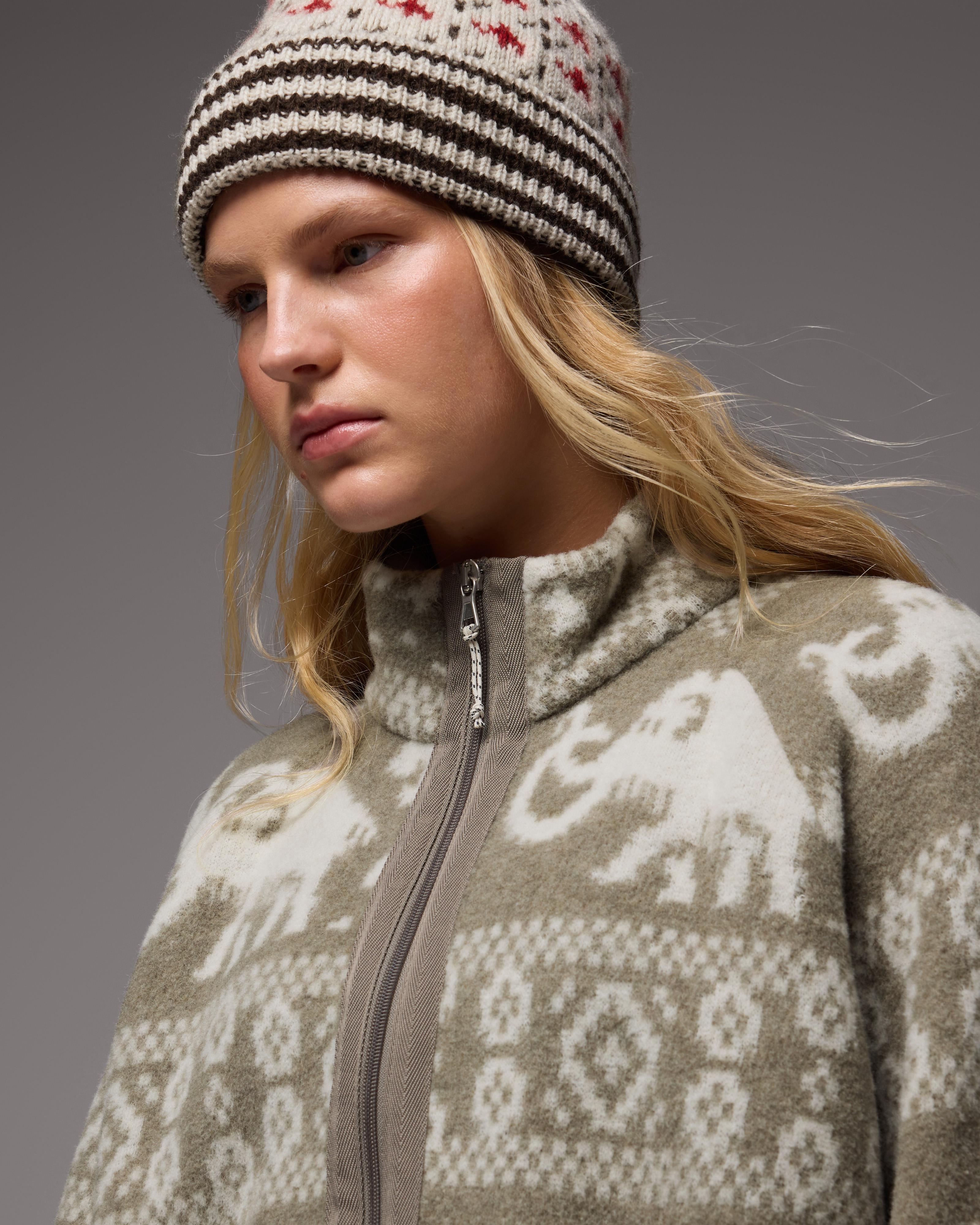 Woman wearing a Mammut-patterned knit hat and zip-up Mammut sweater with white animal motifs, looking down thoughtfully.