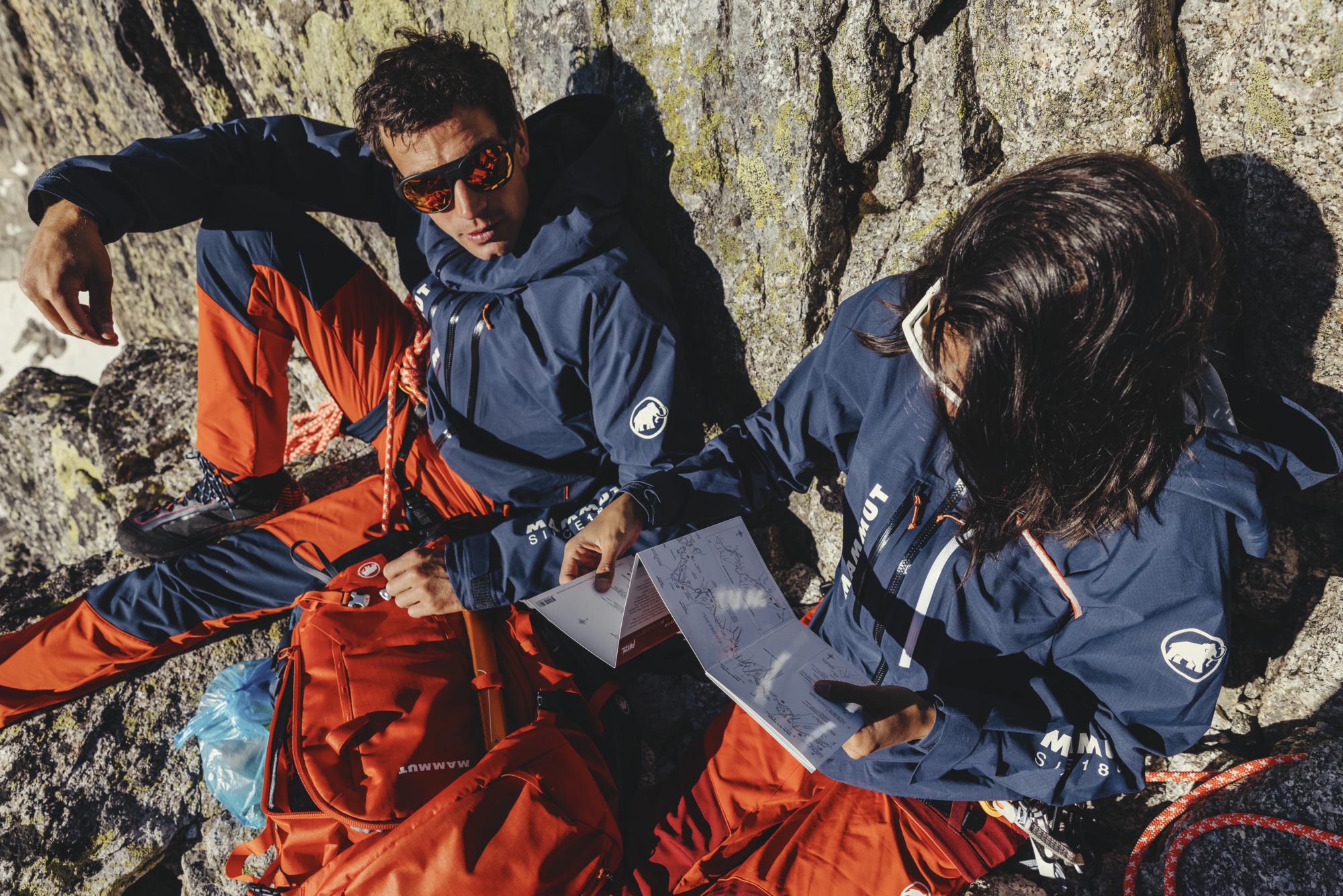 Two mountaineers in blue Mammut jackets and red Mammut pants study a map while sitting on rugged rocky terrain, surrounded by high-quality climbing gear by Mammut.