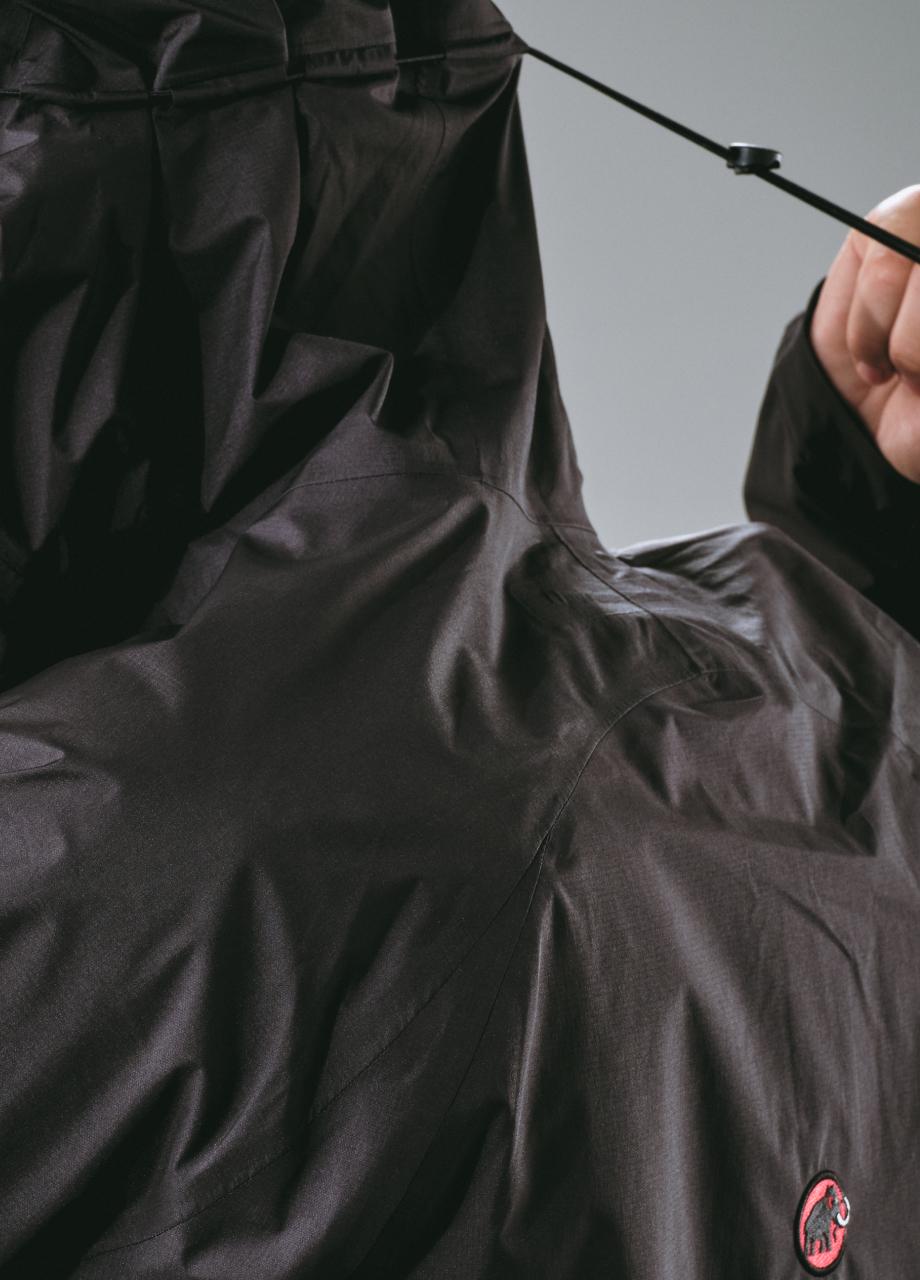 Close-up of a person tightening the hood drawstring on a black Mammut outdoor jacket featuring the brand’s signature red mammoth logo.