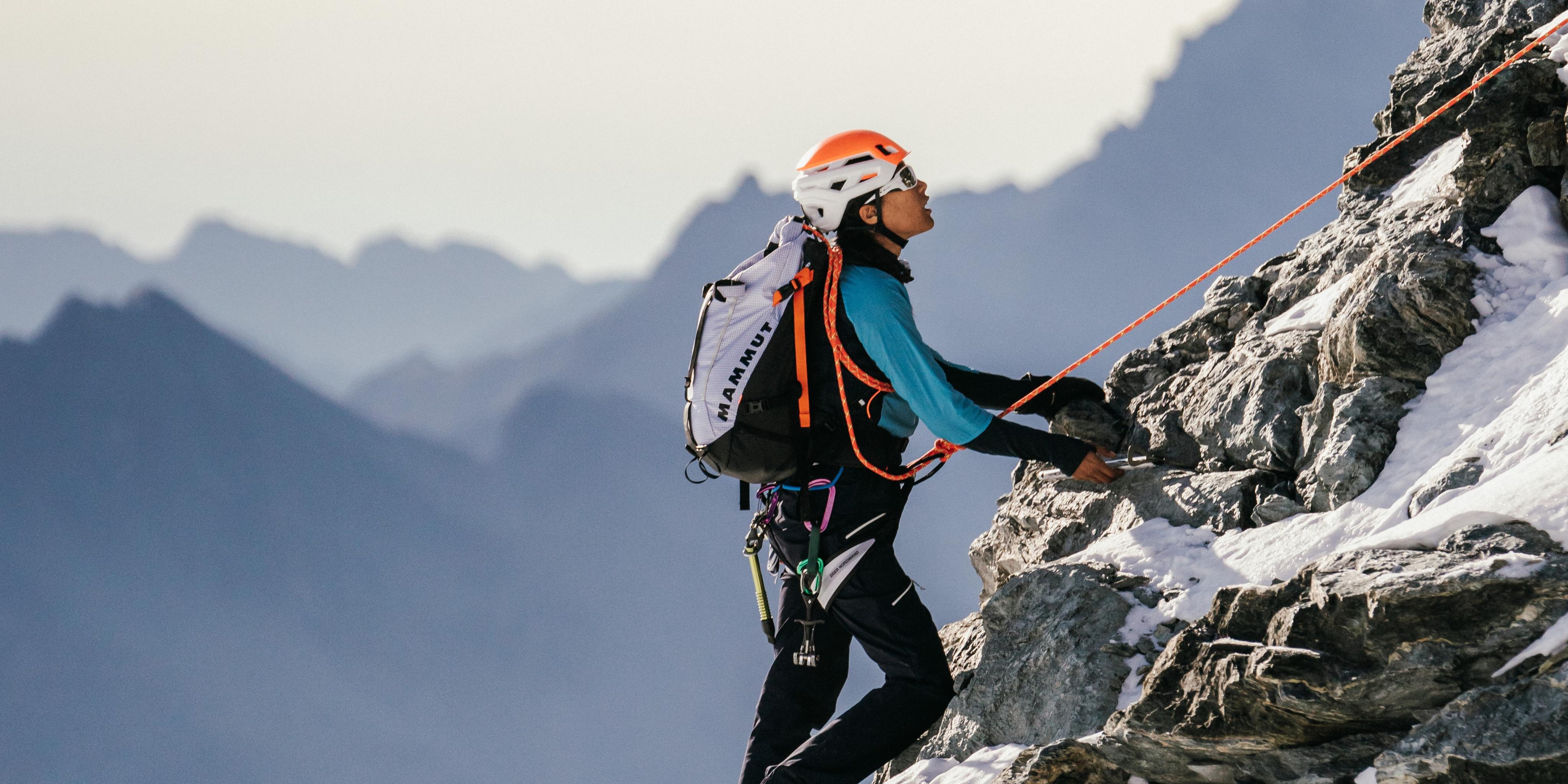 A climber wearing a Mammut orange helmet and blue jacket ascends a steep rocky mountain, secured by a Mammut rope, with majestic distant peaks in the background.