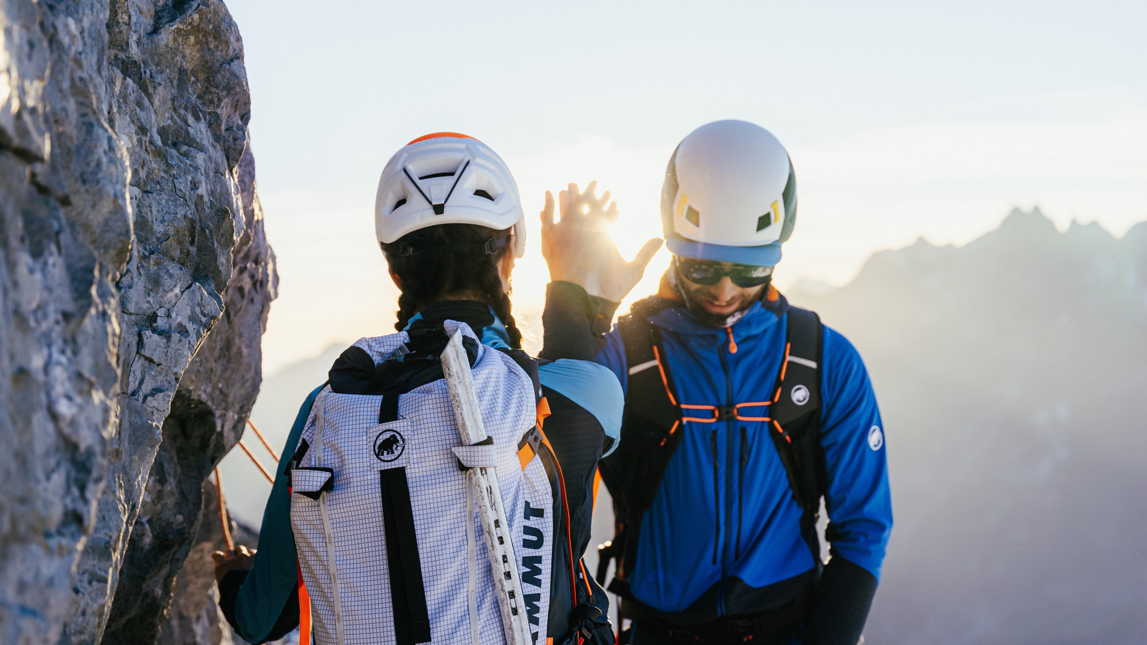 Two climbers in Mammut helmets and gear high-five while scaling a mountain peak at sunrise.