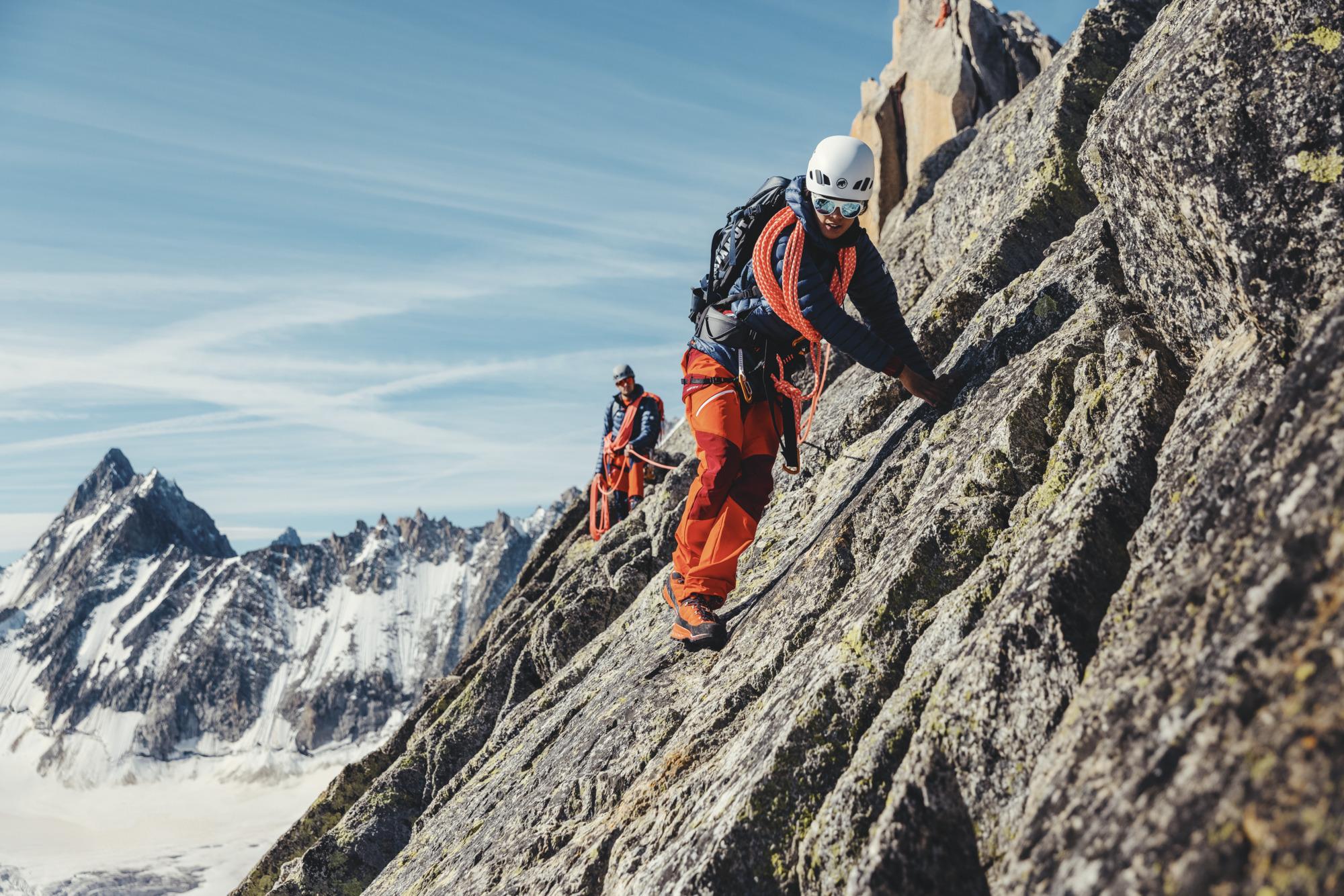 Two climbers, equipped with helmets and Mammut red gear, ascend a rugged rocky mountain with snowy peaks under a clear blue sky in the background.