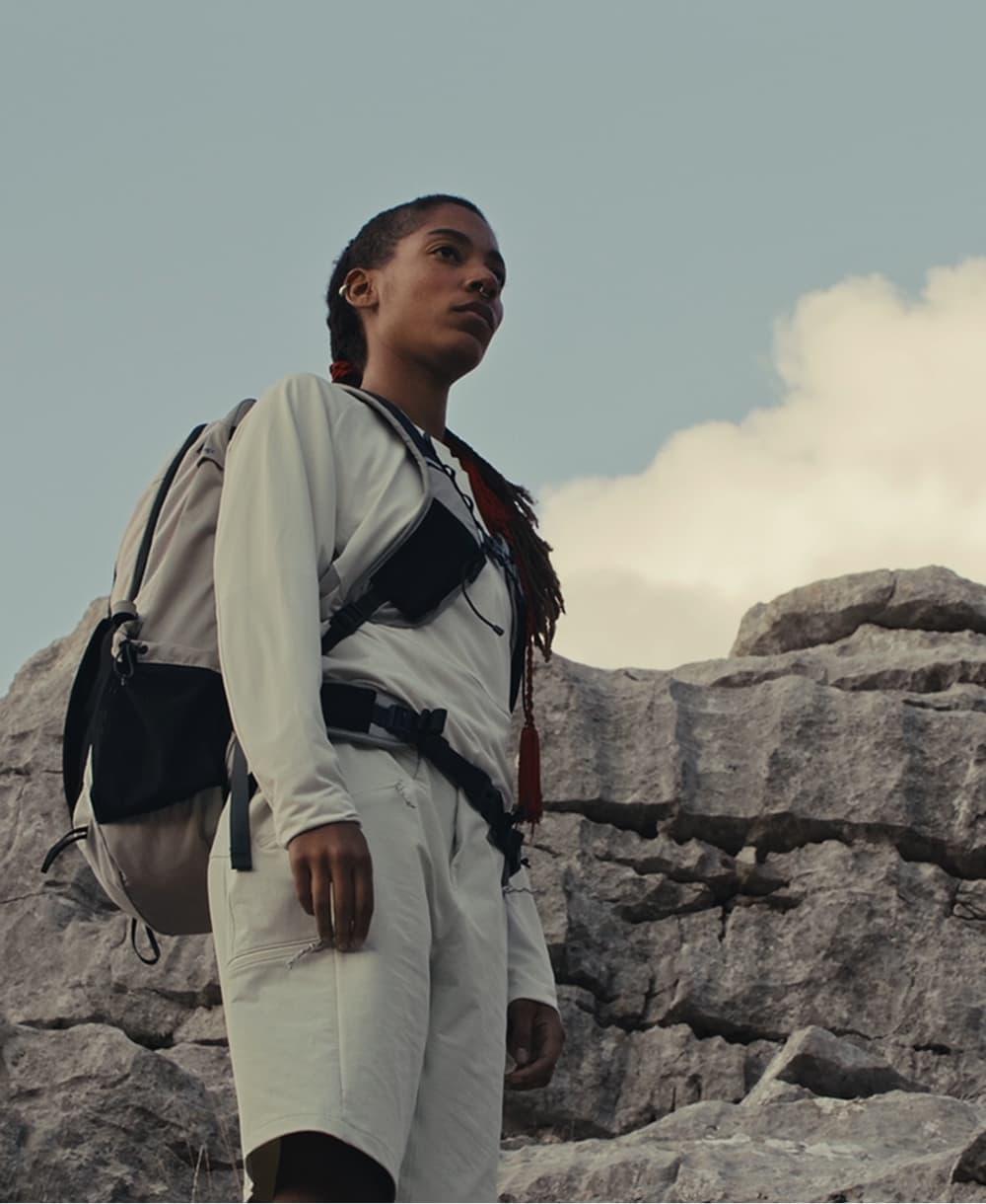 A person in Mammut hiking gear stands on rugged rocky terrain, gazing ahead under a dramatic cloudy sky in the mountains.