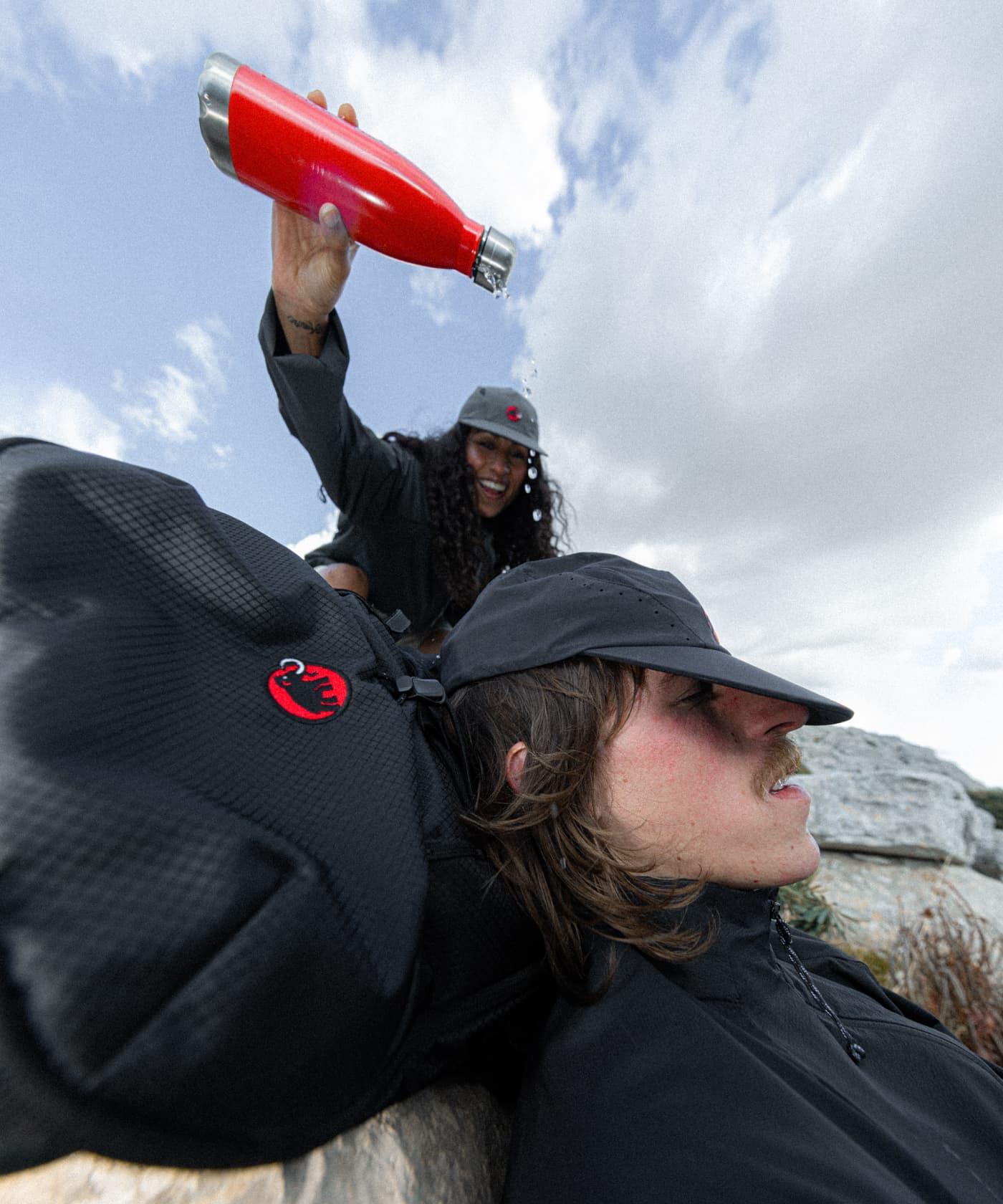 Smiling woman in Mammut gear playfully pours water on a man resting outdoors with a Mammut backpack and hat under cloudy skies.