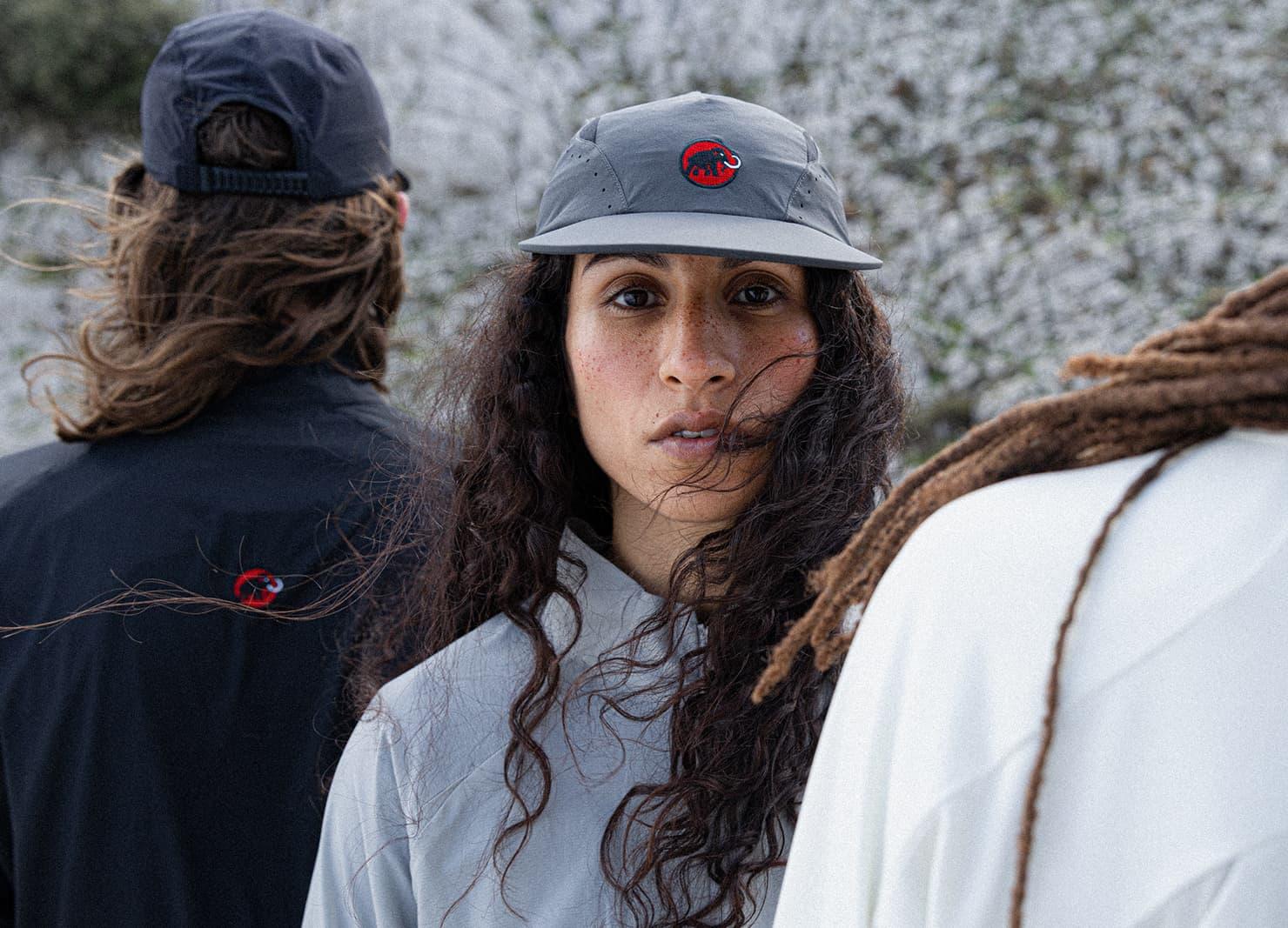 A woman in a gray Mammut cap and jacket stands outdoors between two people, all dressed in outdoor gear with red Mammut logos.