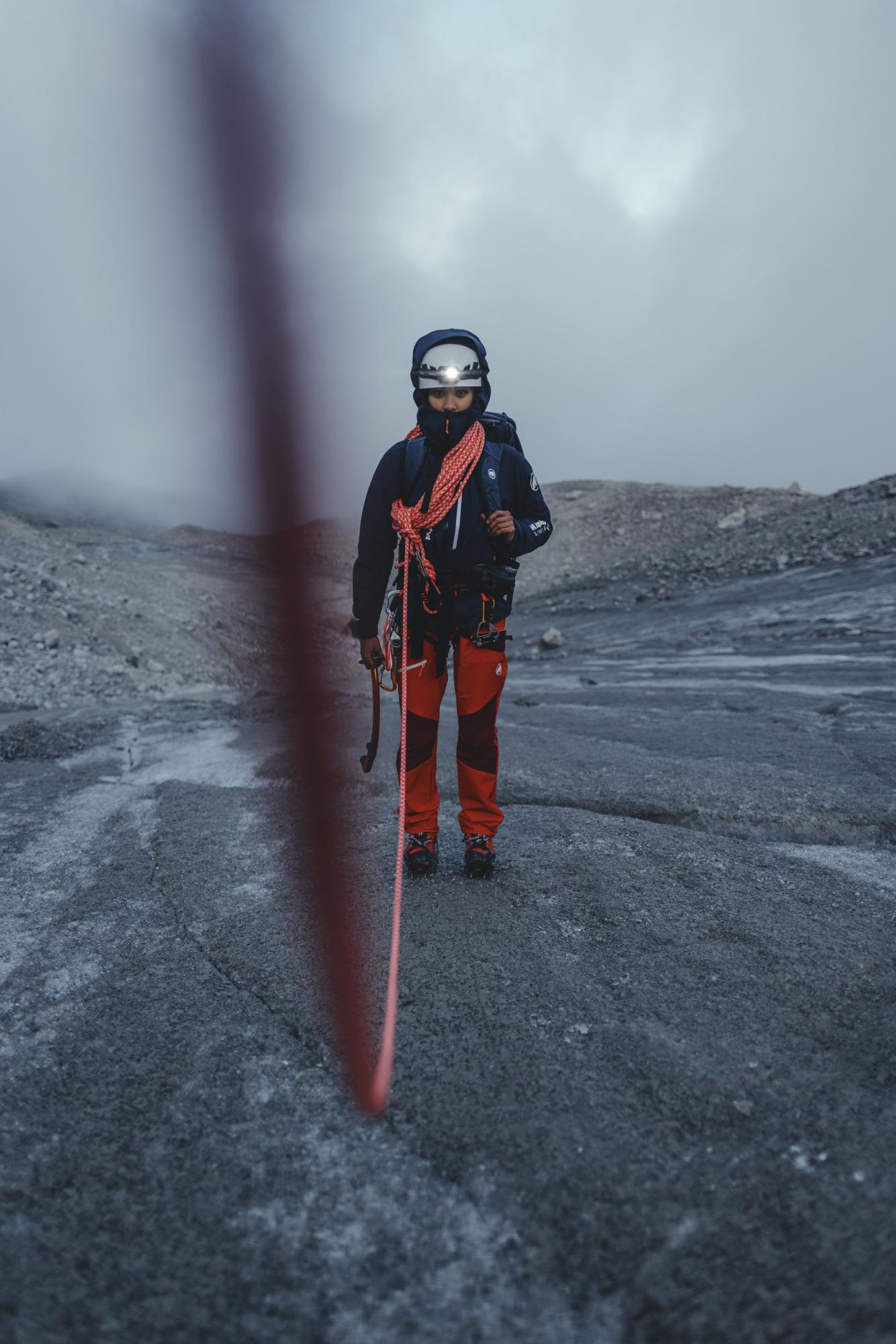 Mammut mountain climber in red and black gear standing on rocky terrain, roped for safety with foggy mountain backdrop.