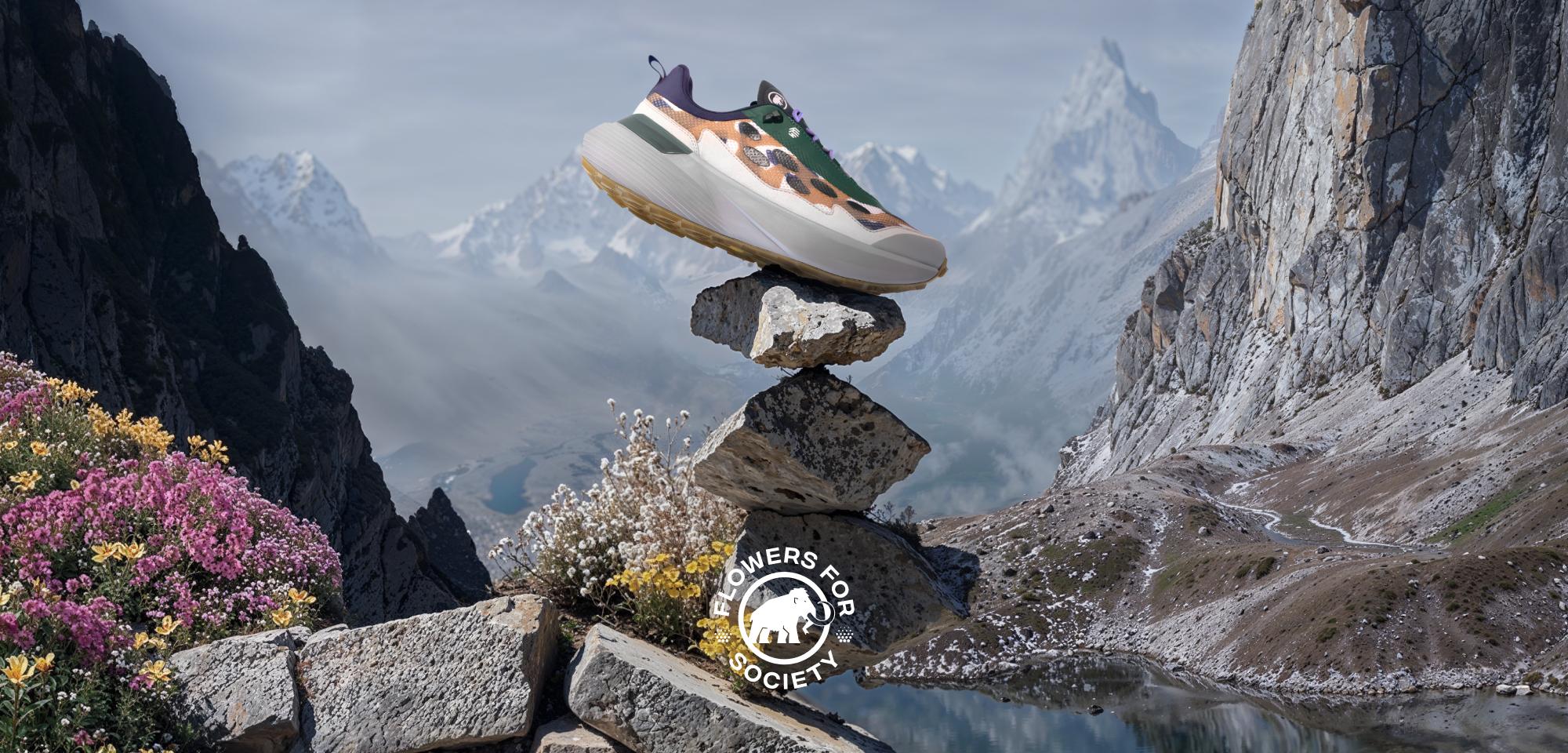 A Mammut sneaker displayed on stacked rocks in a picturesque mountain landscape, surrounded by alpine flowers, rocky cliffs, and distant peaks.