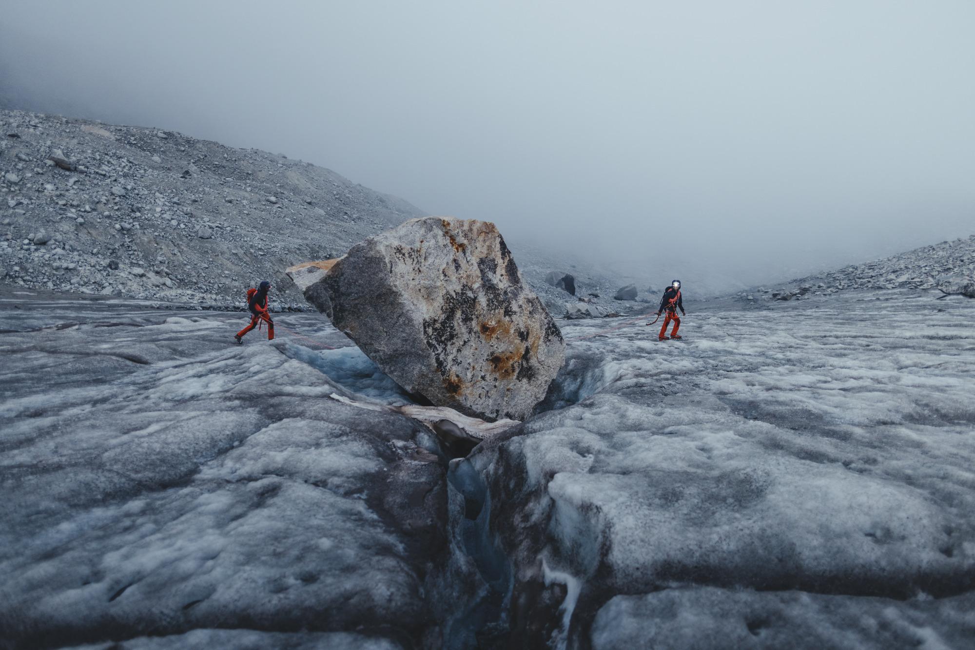 Two climbers in red Mammut gear traverse a glacier, navigating around a prominent boulder with misty alpine scenery in the background.