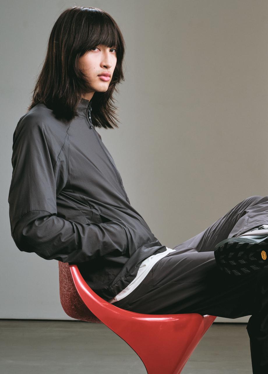 Person with long dark hair in a Mammut gray tracksuit sits sideways on a red chair, looking slightly off camera.
