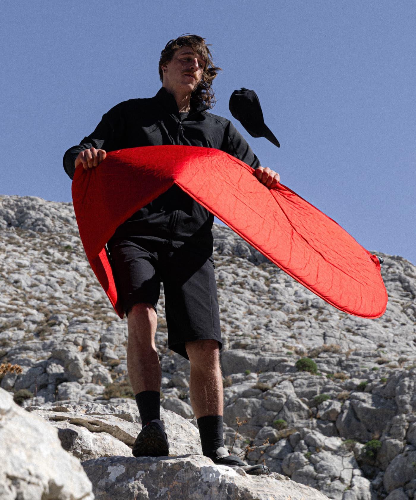 Man standing on rocky terrain holding a bright red Mammut foldable jacket; black cap seen flying off his head in the wind.