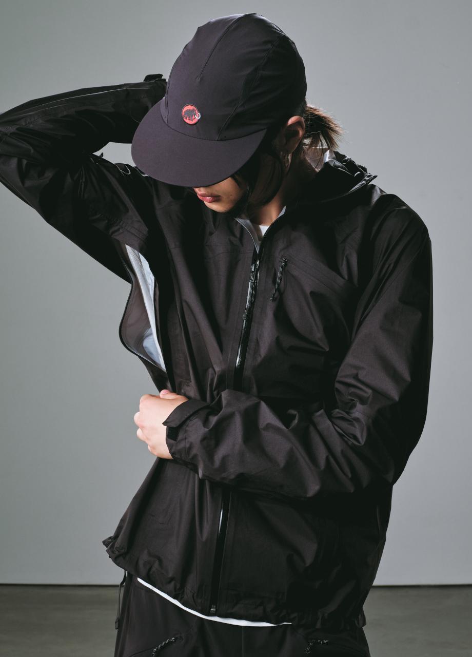 Person in a Mammut black jacket and cap unzipping the jacket’s side vent, facing slightly downward.