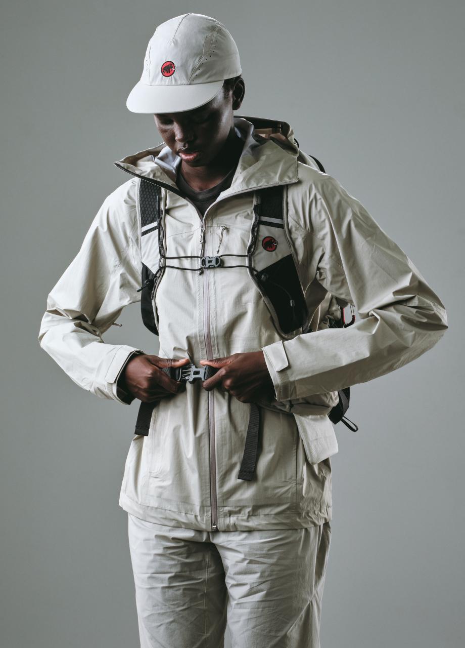 Person wearing a light beige Mammut jacket, pants, cap, and hiking backpack adjusts the chest strap before outdoor activity.