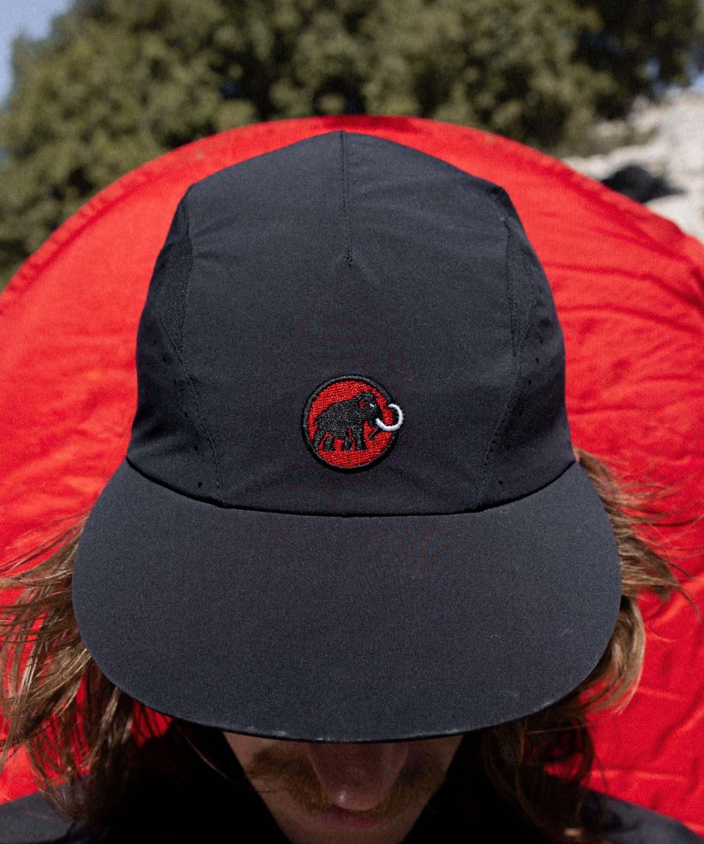 Individual wearing a black Mammut cap with a red mammoth logo, posed before a vivid red circular backdrop.