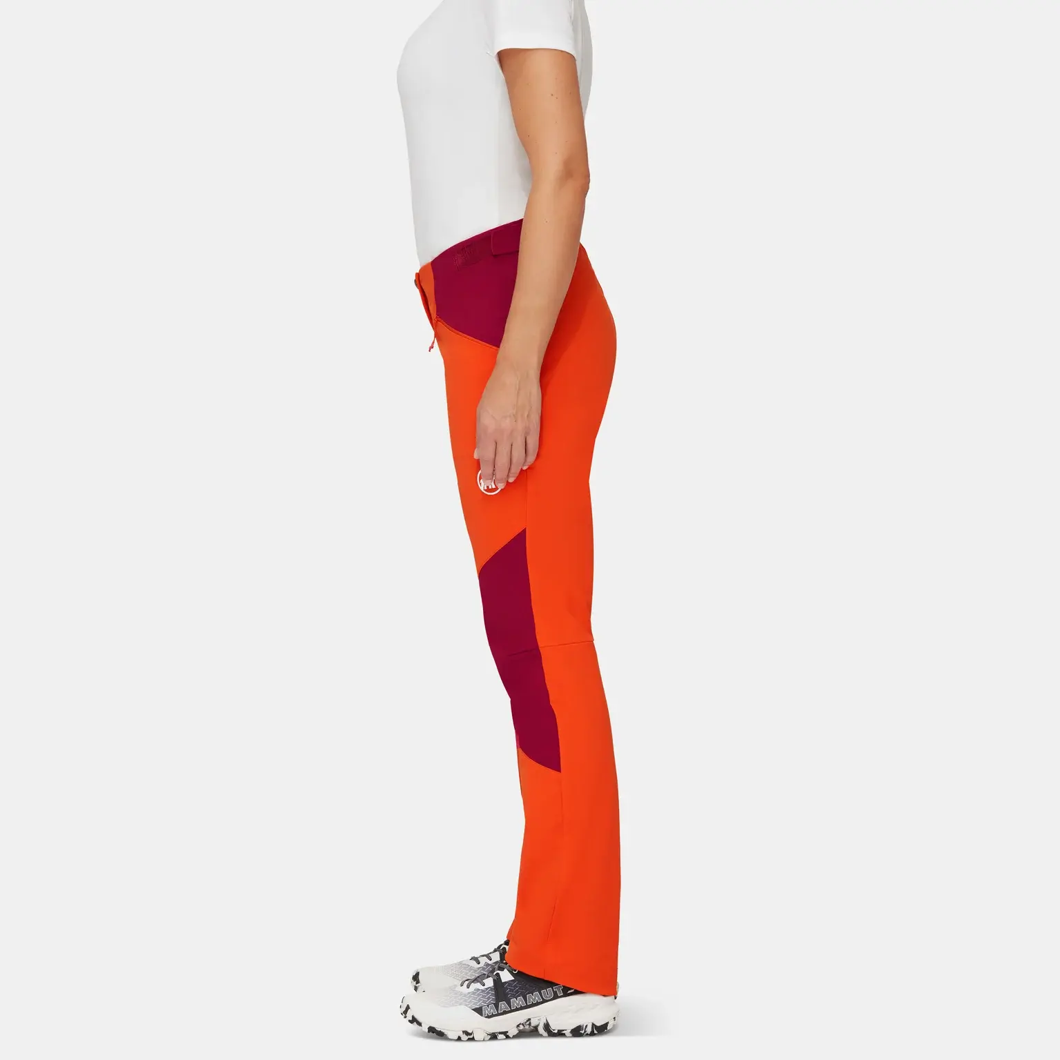 A person wearing Mammut orange and maroon mountaineering pants, a white performance shirt, and white sneakers standing sideways against a white background.