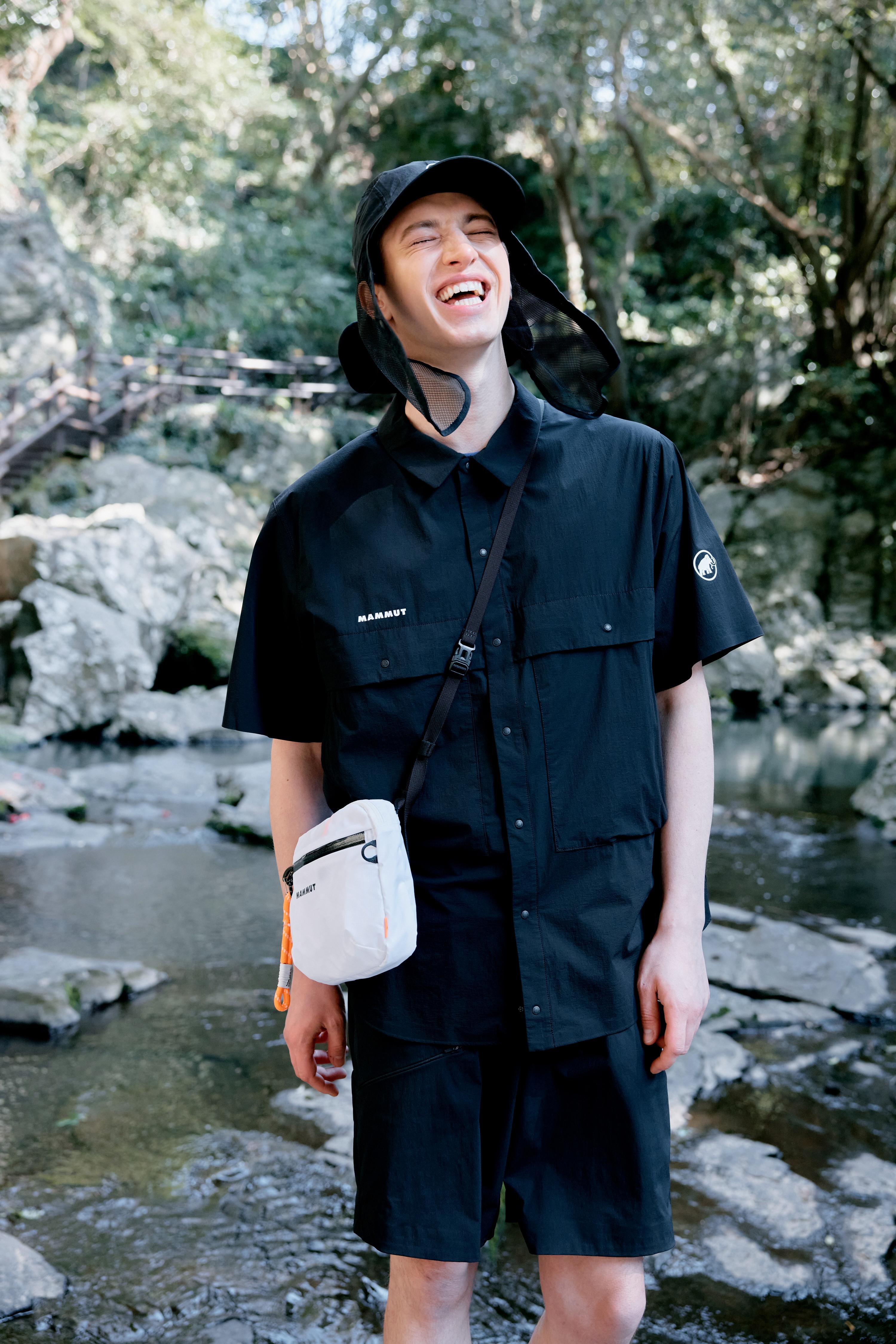 A man in black Mammut outdoor clothing and hat smiles while standing by a rocky stream in a forest.