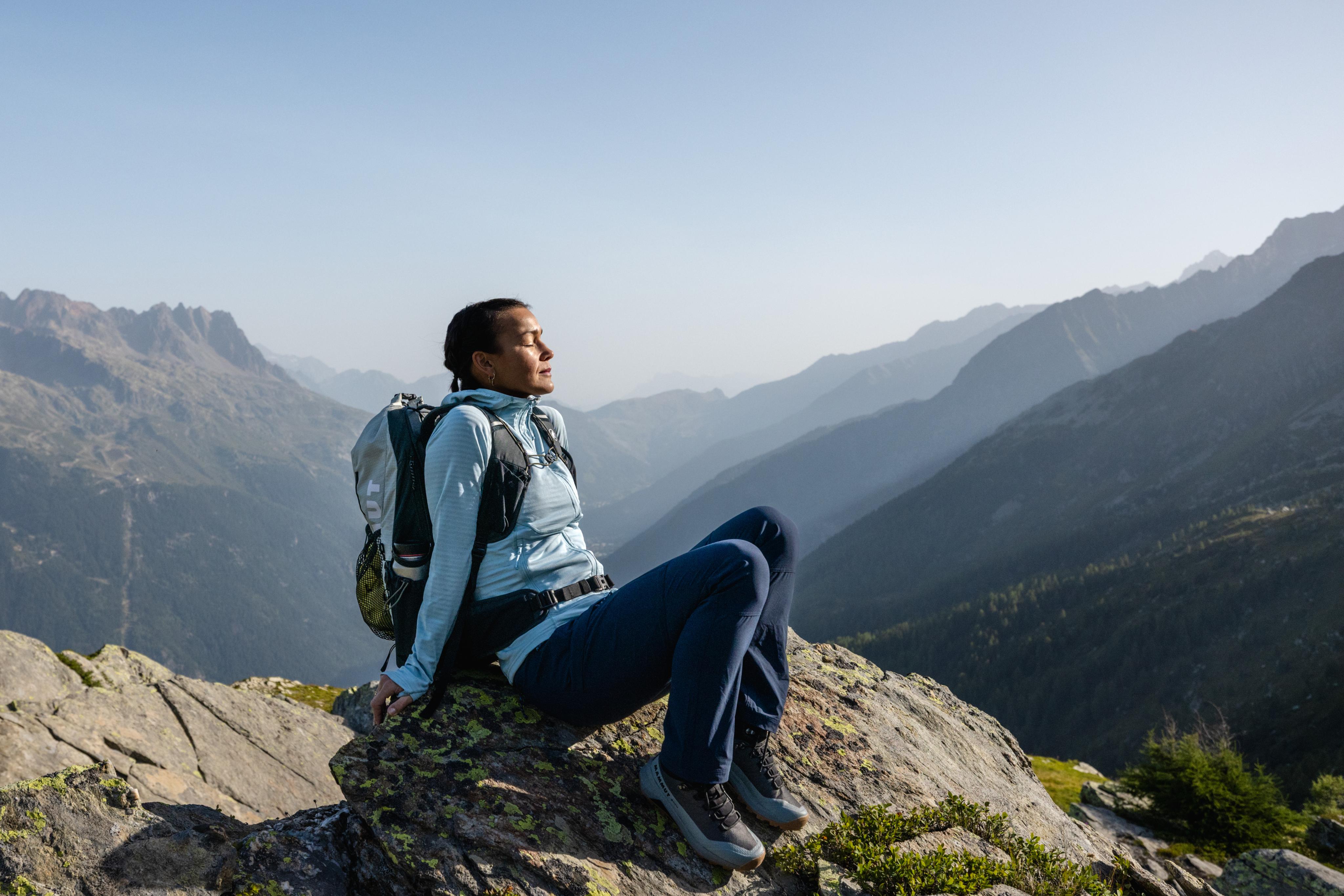 Person in Mammut hiking gear and backpack sits on a rock, enjoying panoramic mountain views and sunlight during an outdoor adventure.