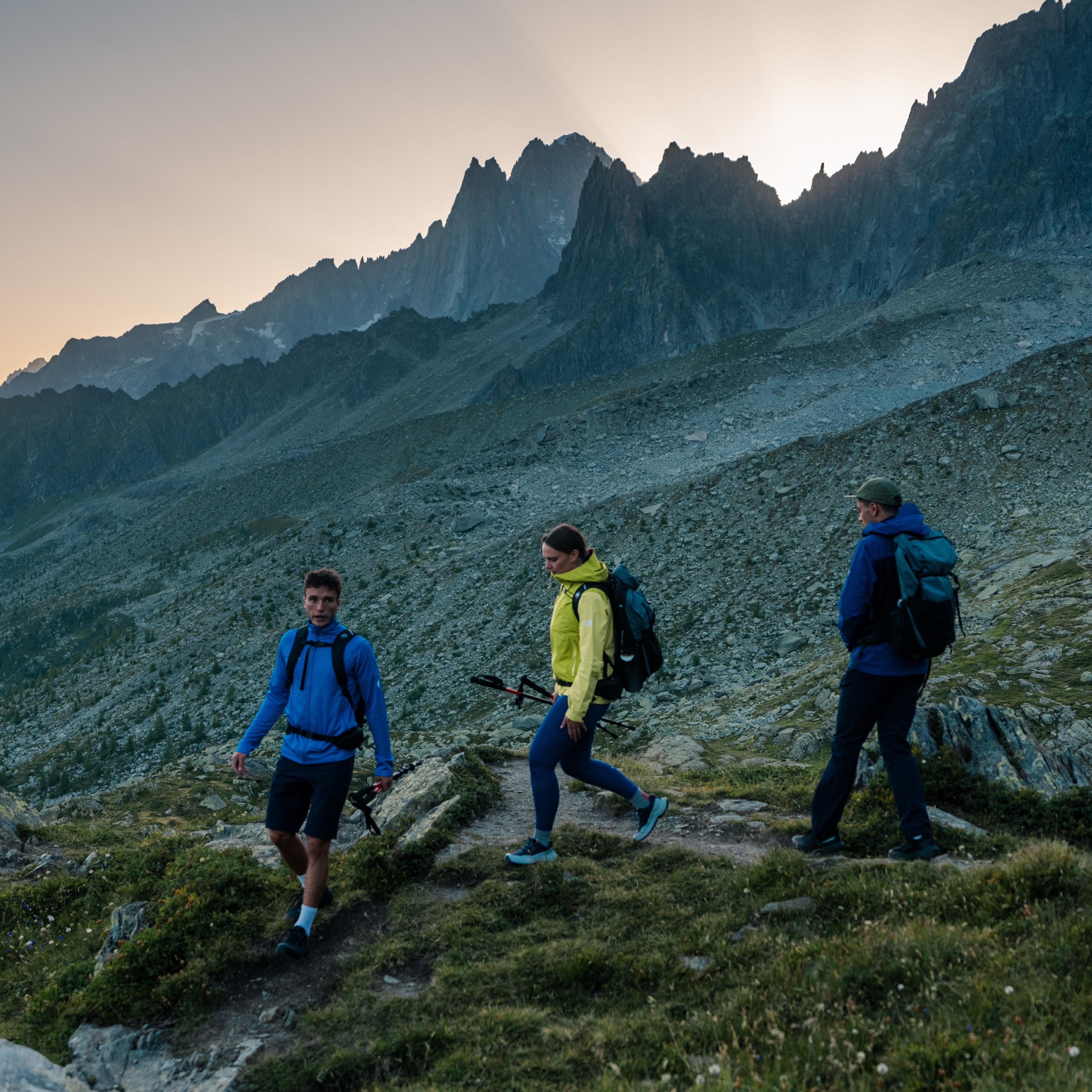 Three hikers in Mammut gear trek a mountain trail at sunrise, surrounded by rugged rocky peaks and alpine scenery in the background.