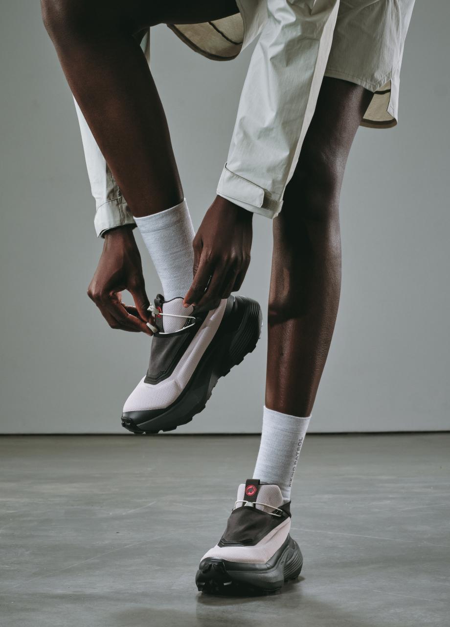 A person in white socks adjusts a strap on Mammut hiking sneakers while wearing a light outdoor Mammut jacket.