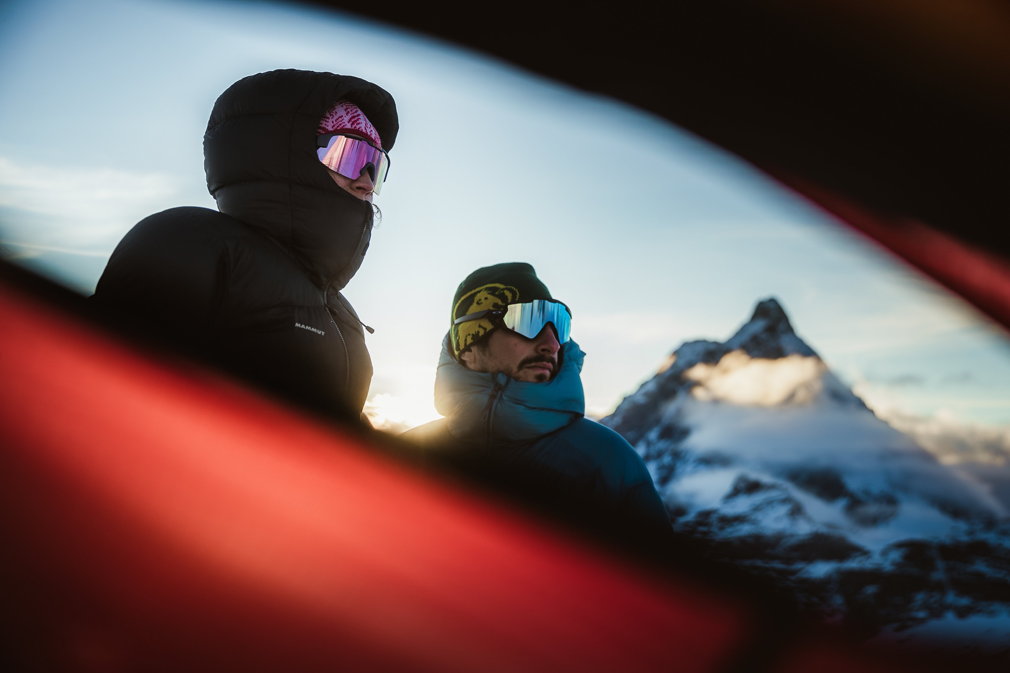 Two mountaineers in Mammut winter gear and reflective sunglasses stand outdoors with a snowy mountain peak in the background.