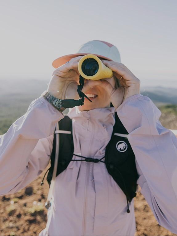 Person wearing a Mammut light jacket using binoculars in the mountains, with clear blue sky in the background.
