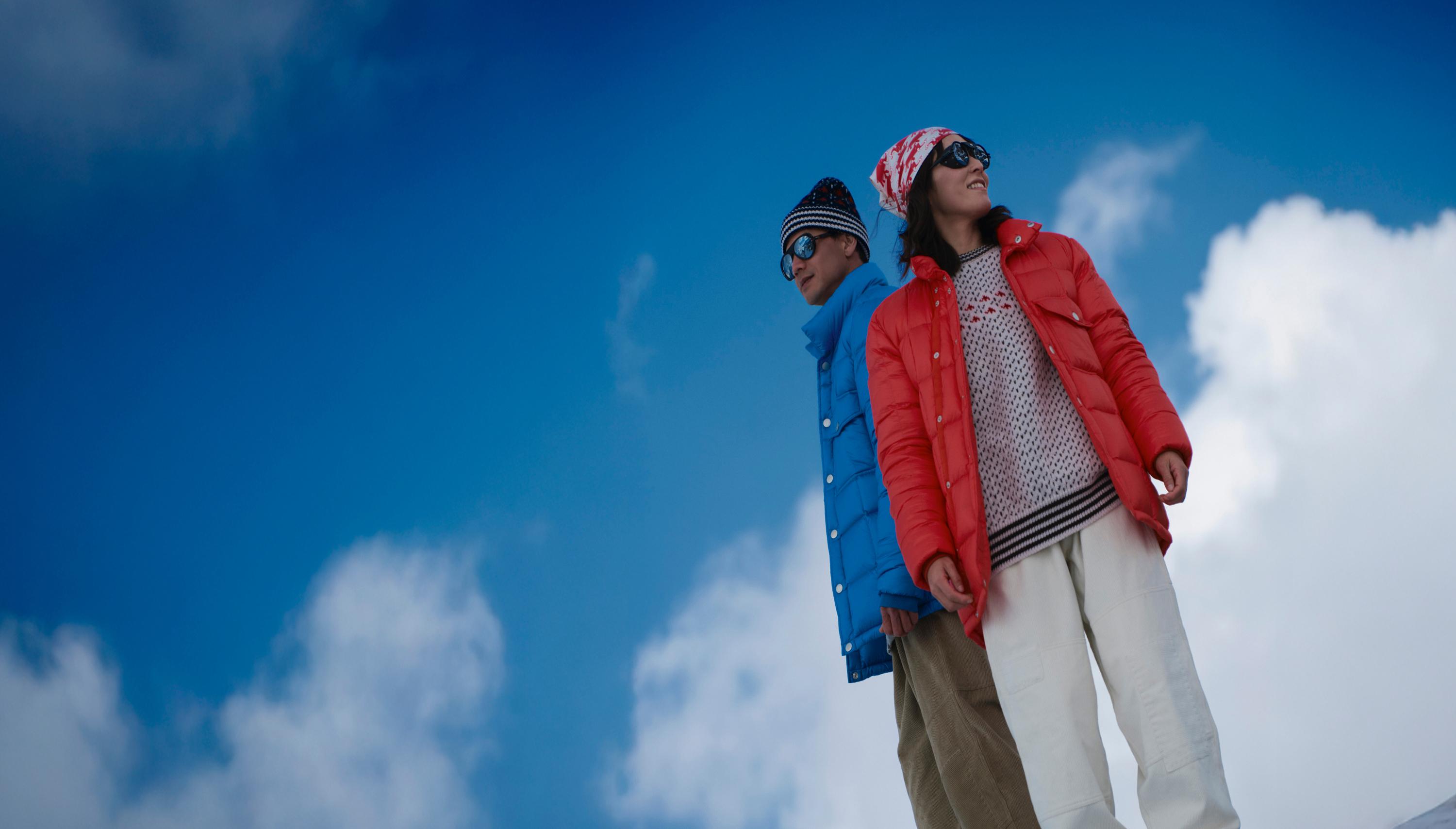 Two people wearing Mammut winter jackets and sunglasses stand outdoors against a blue sky with scattered clouds.