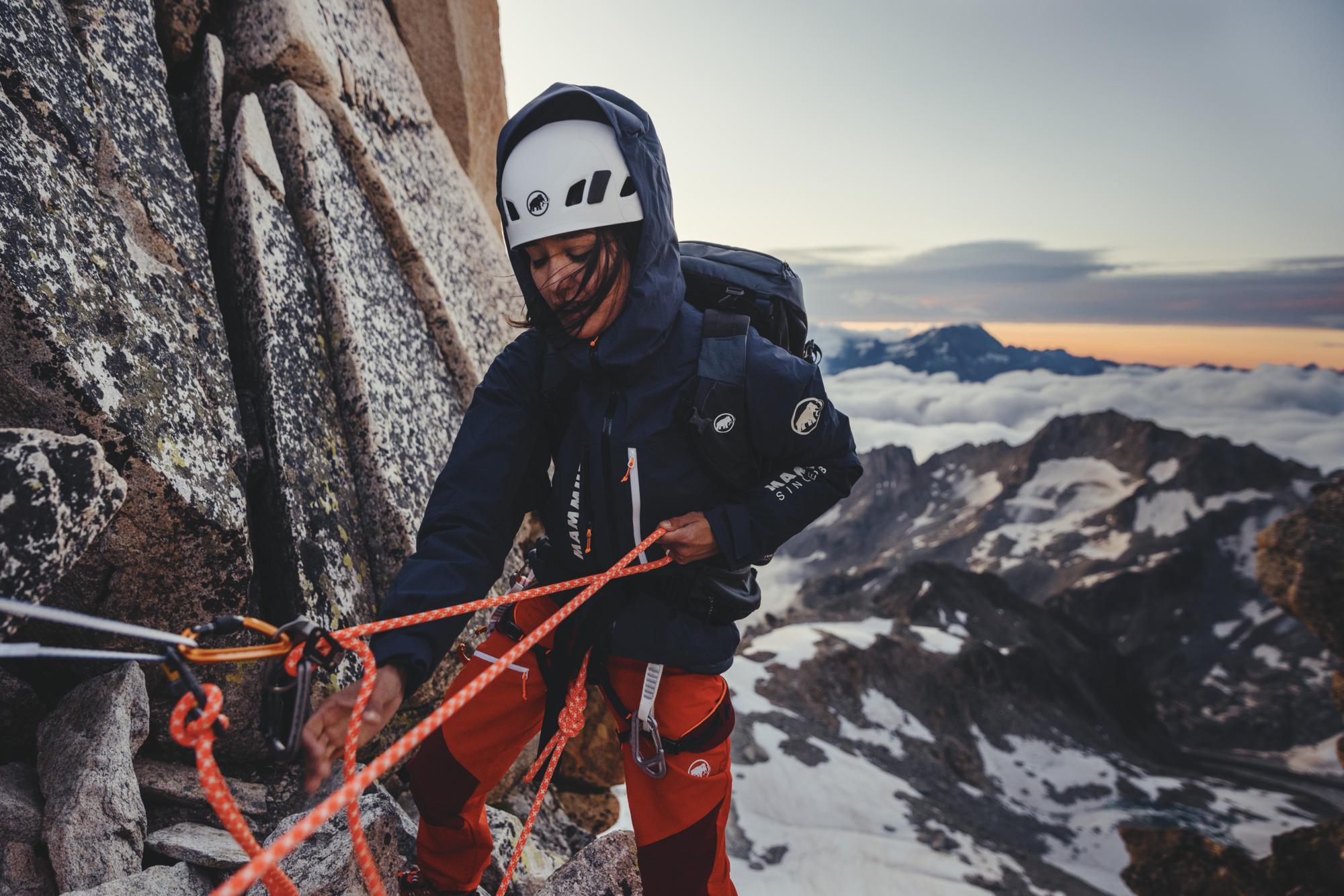 A climber wearing a Mammut helmet adjusts ropes on a mountain, framed by a scenic snowy peak and cloudy sky backdrop.