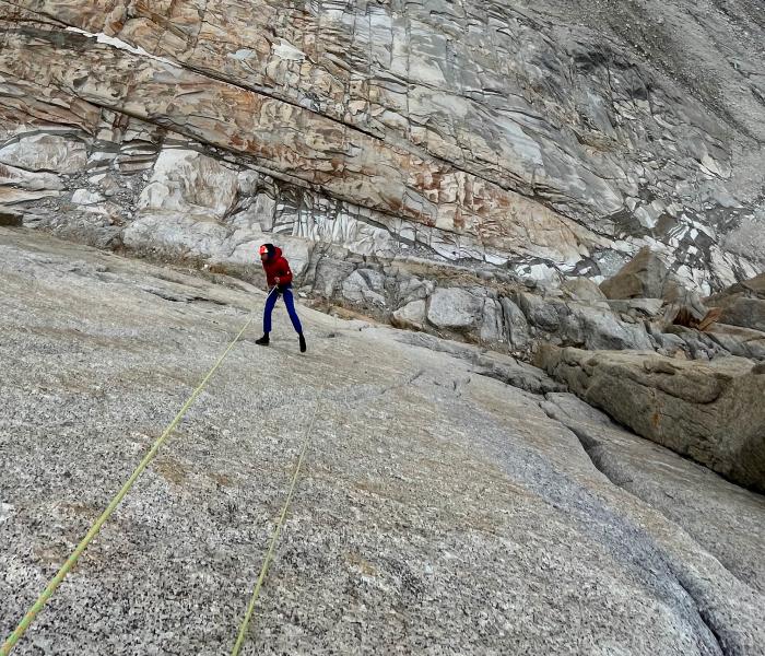 A climber in a red Mammut jacket and blue pants rappelling down a steep, rocky cliff with Mammut ropes.