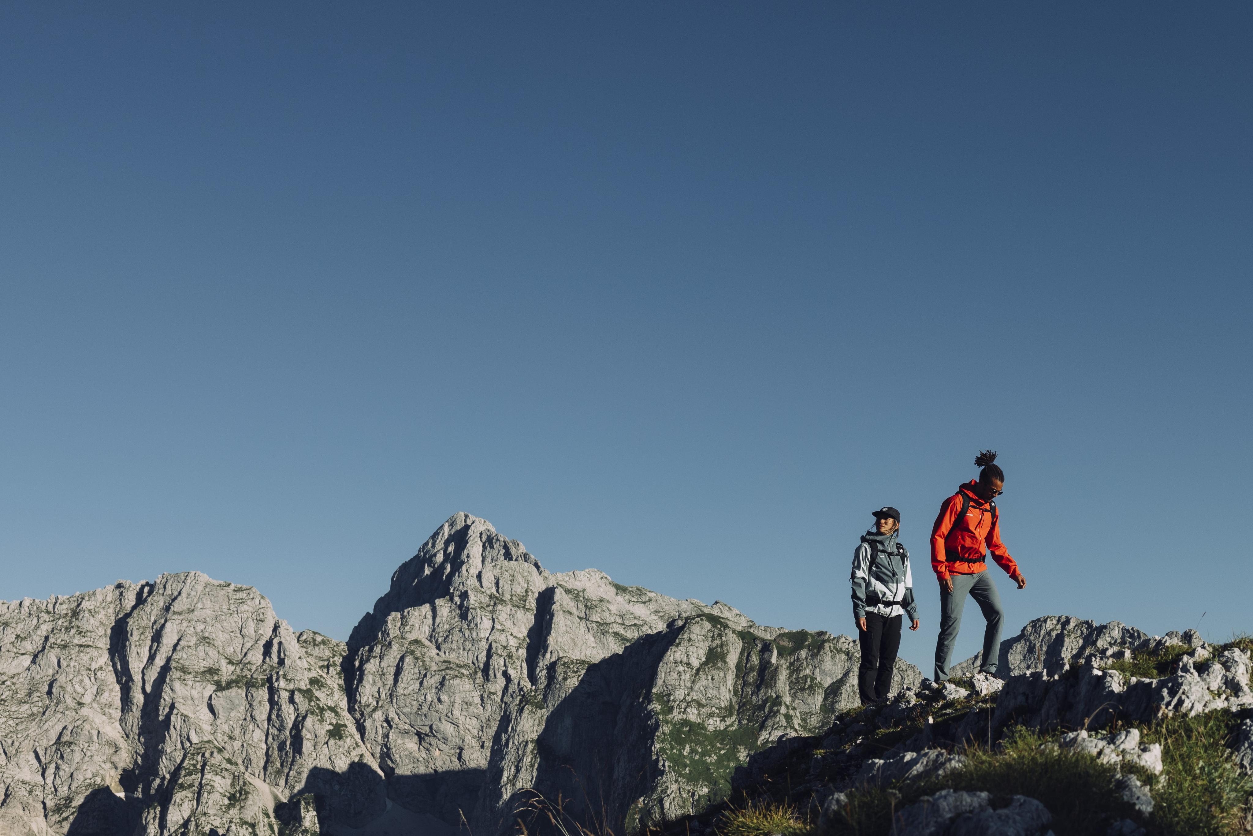 Two hikers in Mammut gear trek a rugged mountain ridge, towering peaks and a clear blue sky creating a dramatic alpine backdrop.