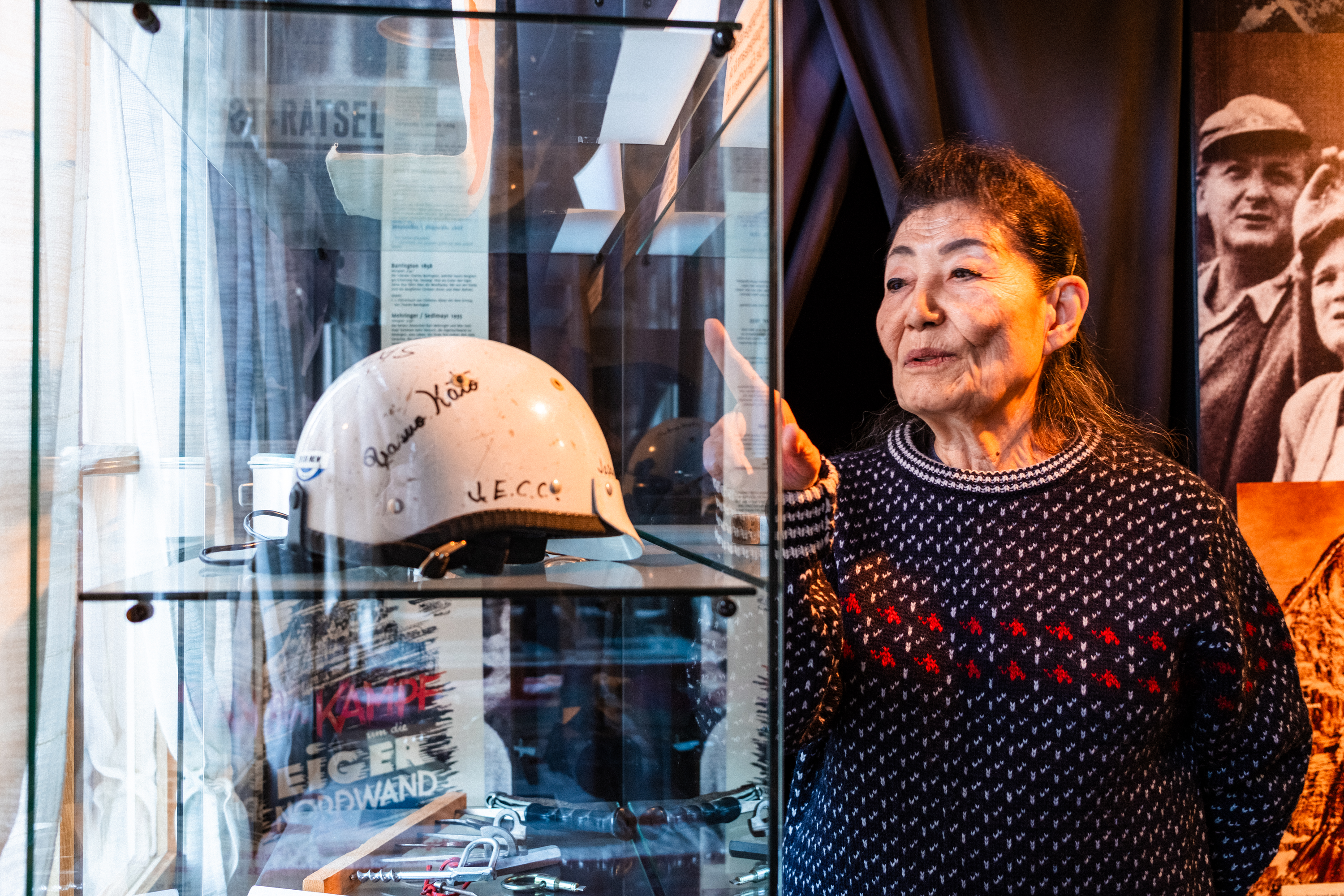 Michiko Imai points at a signed climbing helmet displayed in a glass case at a mountaineering museum exhibit.
