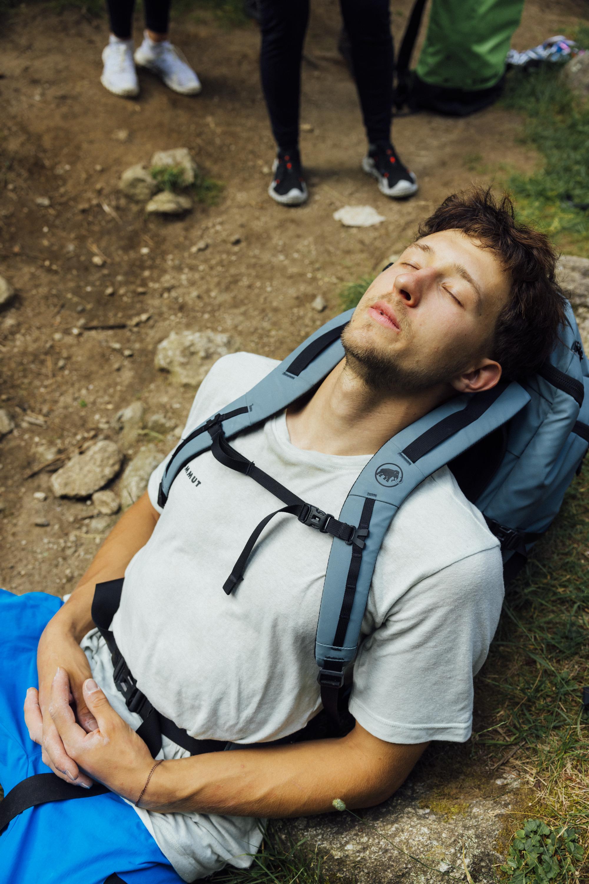 A man in Mammut hiking gear rests on the ground with his eyes closed, using his Mammut backpack as a pillow.