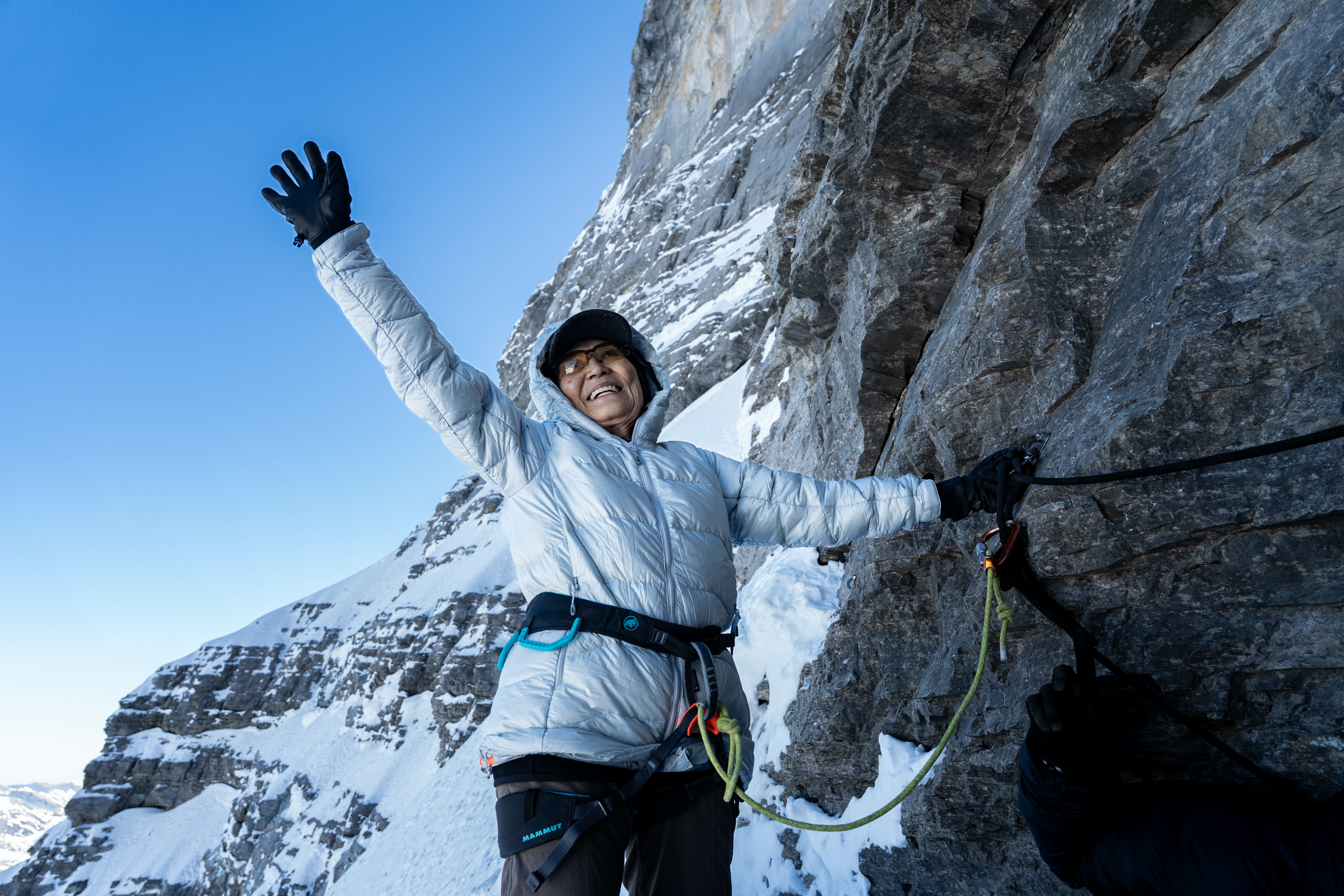 Smiling climber in Mammut winter gear stands on snowy mountain, raising one arm joyfully beside a rugged rocky cliff.
