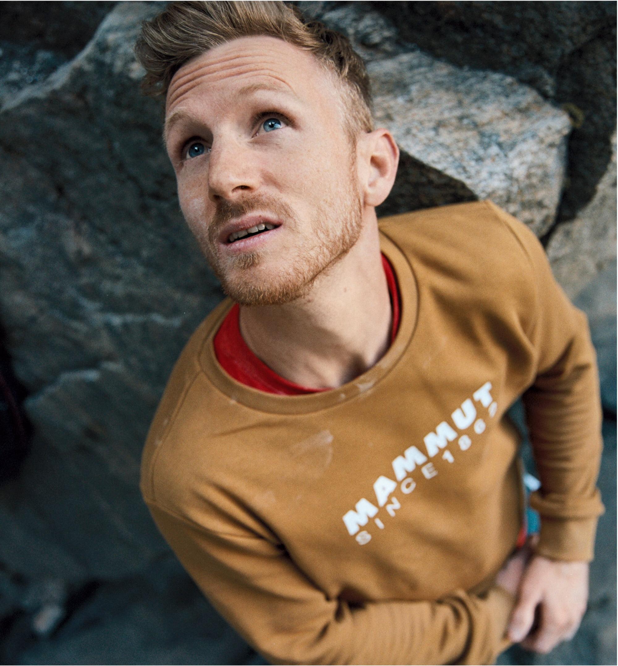 Person wearing a brown Mammut sweatshirt, gazing upwards thoughtfully against a rugged rocky background.